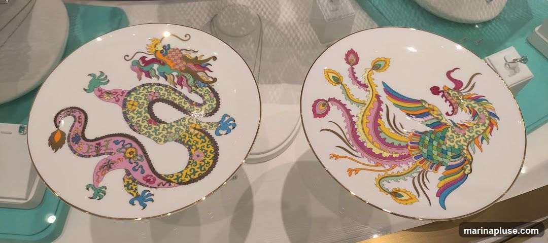 Two decorative white plates with gold rims are displayed. The plate on the left features a colorful dragon design, while the plate on the right showcases a vibrant phoenix design. Both plates have intricate patterns and are presented in a retail display setting, possibly a jewelry or home goods store, with other items visible in the background.