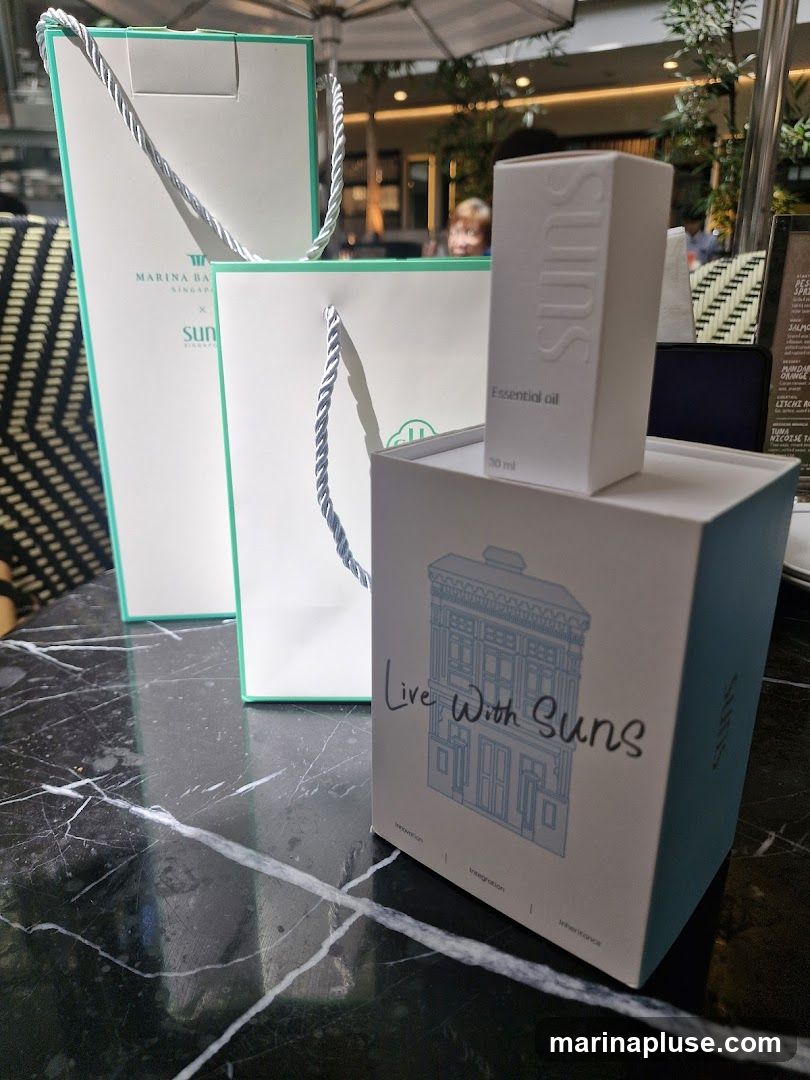 A close-up shot of packaging for "Suns" brand essential oil, featuring two shopping bags and a box with an illustration of a building. The box has the text "Live With Suns" and branding for "Integration" and "Inheritance". A small bottle of essential oil is placed on top of the box.