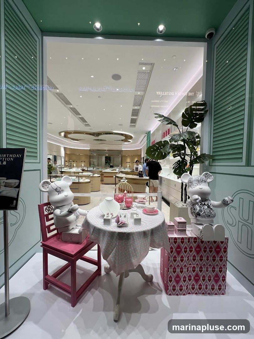 A whimsical display in a jewelry store window features two large bear-like figures, one seated at a small table laden with jewelry and the other standing next to a stack of pink boxes. The table is draped with a floral cloth and set with a necklace, earrings, and rings. The background shows the interior of the store with display cases and customers. A sign on the left advertises a "Birthday Option."