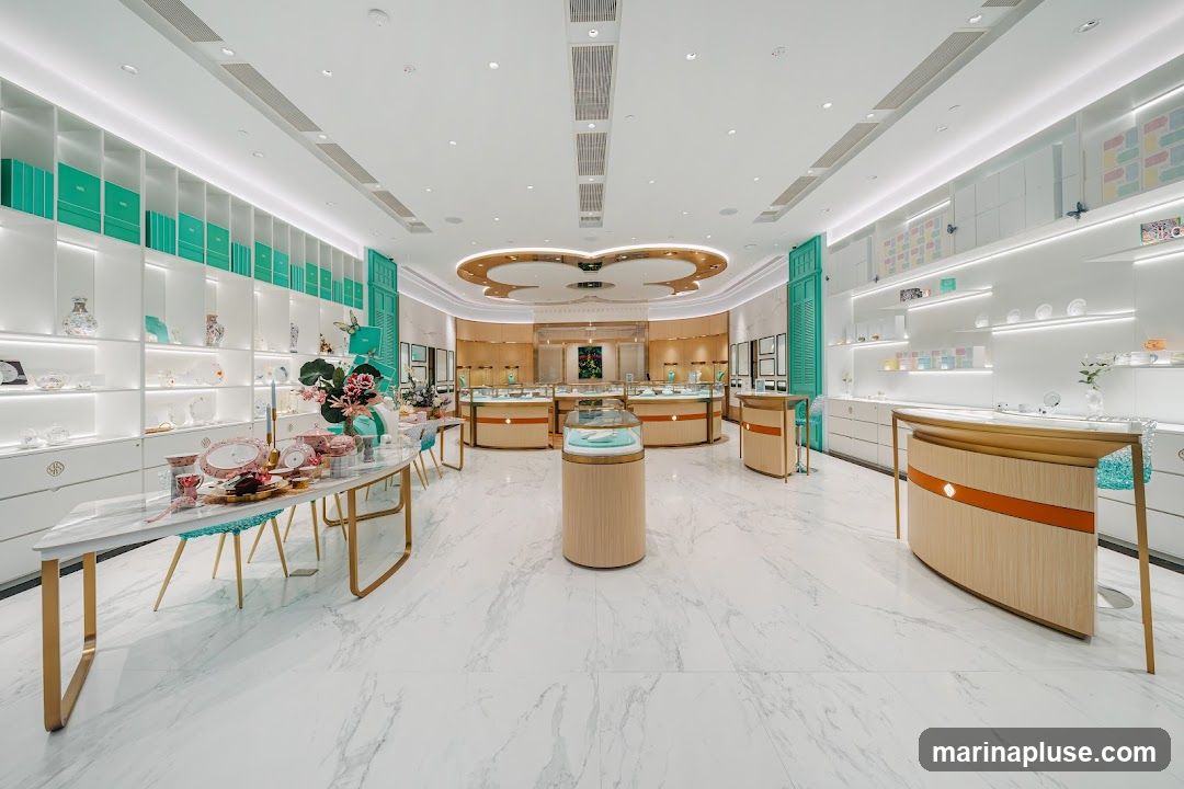 A brightly lit, modern jewelry store interior with white marble floors. Shelving units line the walls, displaying various items including decorative boxes, vases, and tableware. A long display table in the foreground showcases more decorative items and floral arrangements. In the center of the store, several display cases are arranged, with a prominent circular display case in the foreground. The overall aesthetic is clean, elegant, and luxurious.