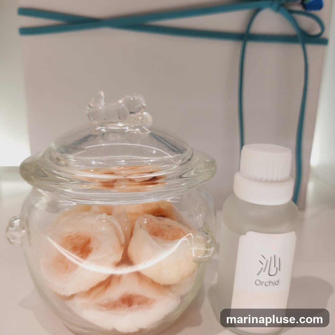 A glass jar filled with what appears to be dried fruit slices, possibly potpourri or a decorative item. Next to it is a small frosted glass bottle with a white cap. The bottle has a label with Japanese characters and the word "Orchid" printed below. In the background, a white gift box is tied with a teal ribbon.