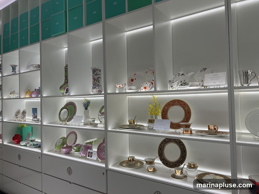 A well-lit display of various porcelain items, including vases, plates, bowls, teacups, and saucers, arranged on shelves within a store. The items feature intricate designs and patterns, with some pieces in pastel colors and others with gold accents. Boxes are stacked on the top shelves.