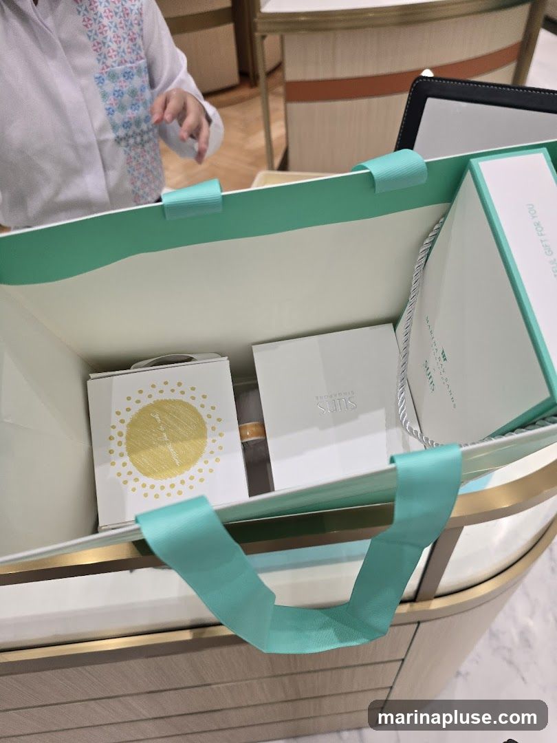 A shopping bag with turquoise handles contains several gift boxes. One box has a yellow sun design with the text "Jewels by my creations". Another box is white with the text "Suns Singapore". A larger gift box on the right has text that reads "A special gift for you". A person wearing a white shirt with a patterned yoke is visible in the background.