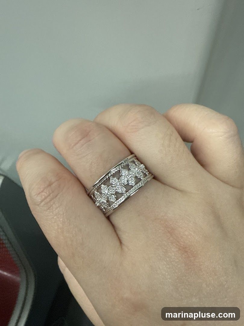 A close-up shot of a hand wearing a wide, ornate ring. The ring is made of white metal and features a delicate floral pattern, with small diamonds set into each flower. The background is a plain, light gray wall.