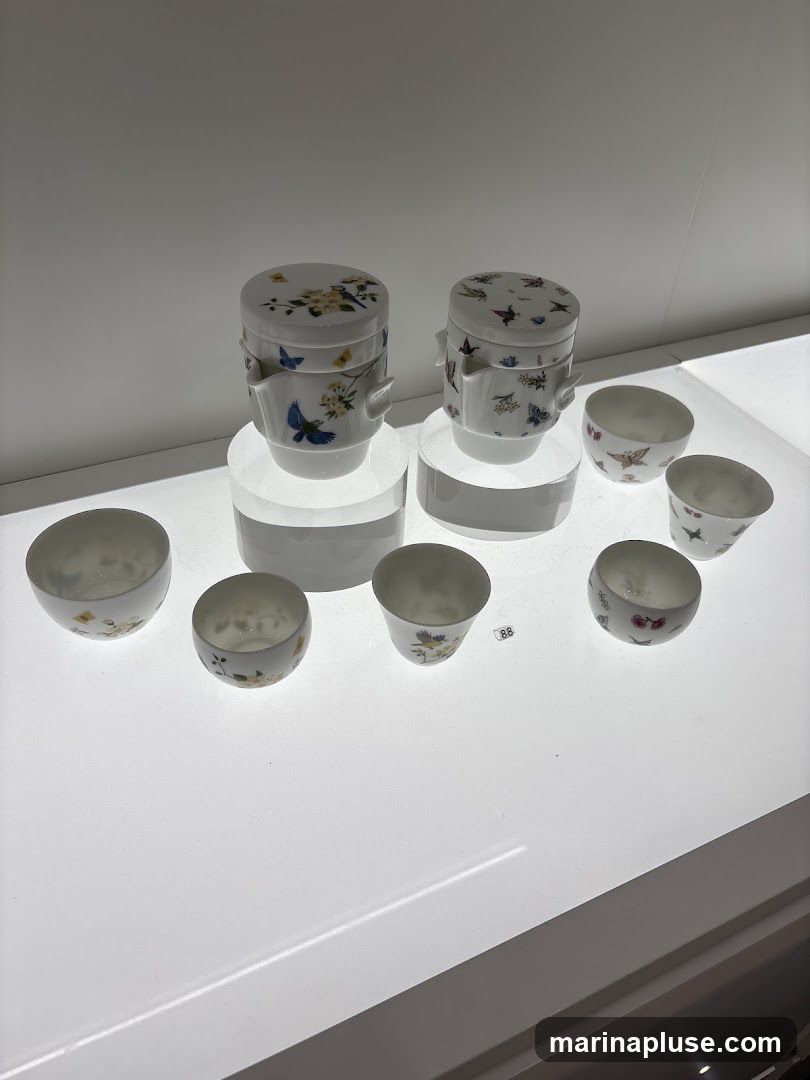 A display of white porcelain teaware, including two lidded teapots and several small cups. The teapots and cups are adorned with delicate hand-painted designs of flowers, birds, and butterflies. The items are arranged on white pedestals under bright lighting, suggesting a retail or exhibition setting.