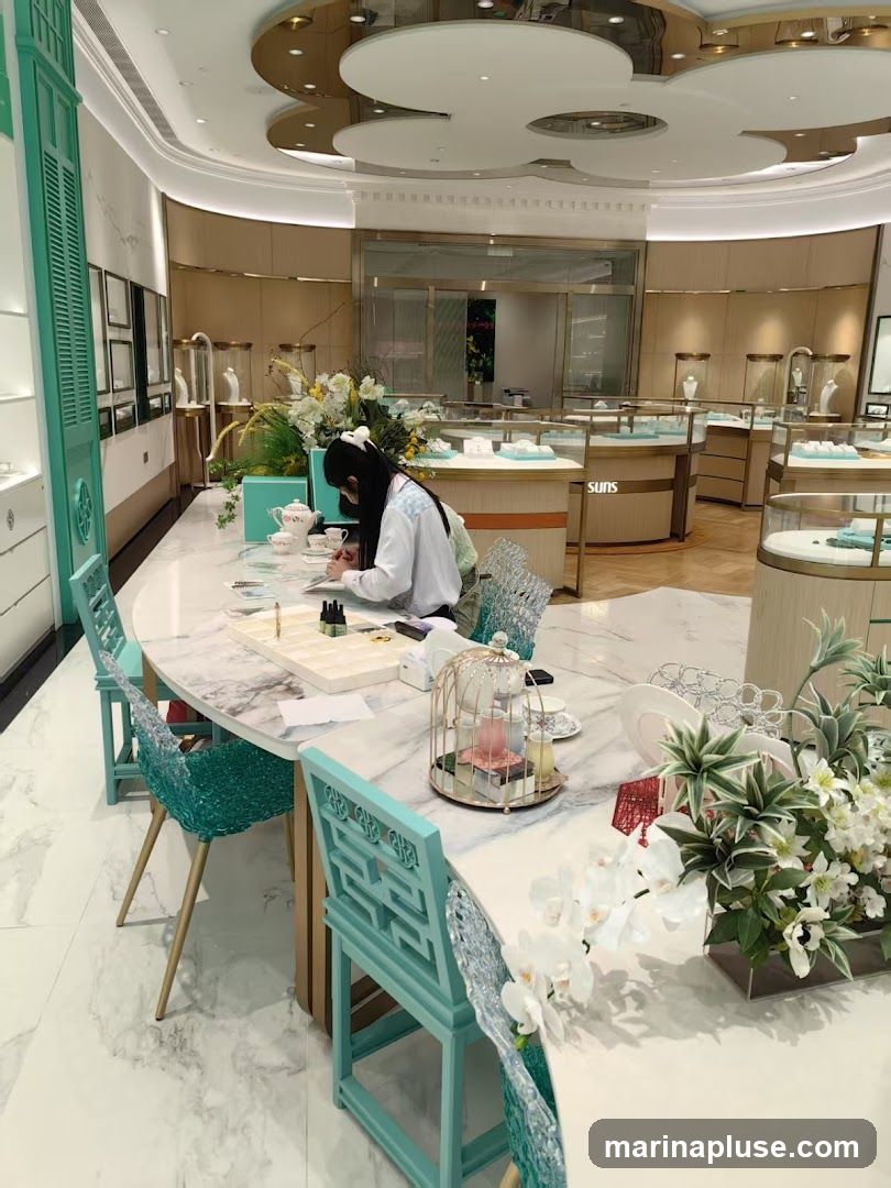 A woman is seated at a marble table in a well-lit jewelry store, writing in a notebook. The table is adorned with a tea set, floral arrangements, and decorative birdcages. Display cases filled with jewelry are visible in the background, along with branded signage that reads "SUNS". The store features elegant decor with a light color palette and ornate ceiling details.