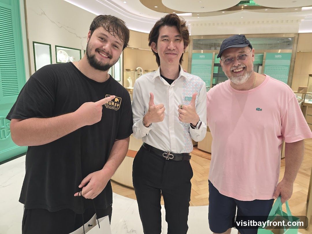 Three men are posing for a photo inside a brightly lit store. The man on the left is wearing a black t-shirt and pointing to his shirt. The man in the middle is wearing a white collared shirt with a patterned design and giving a thumbs up. The man on the right is wearing a pink t-shirt and a baseball cap, also giving a thumbs up. The background shows display cases with jewelry and the store's branding.