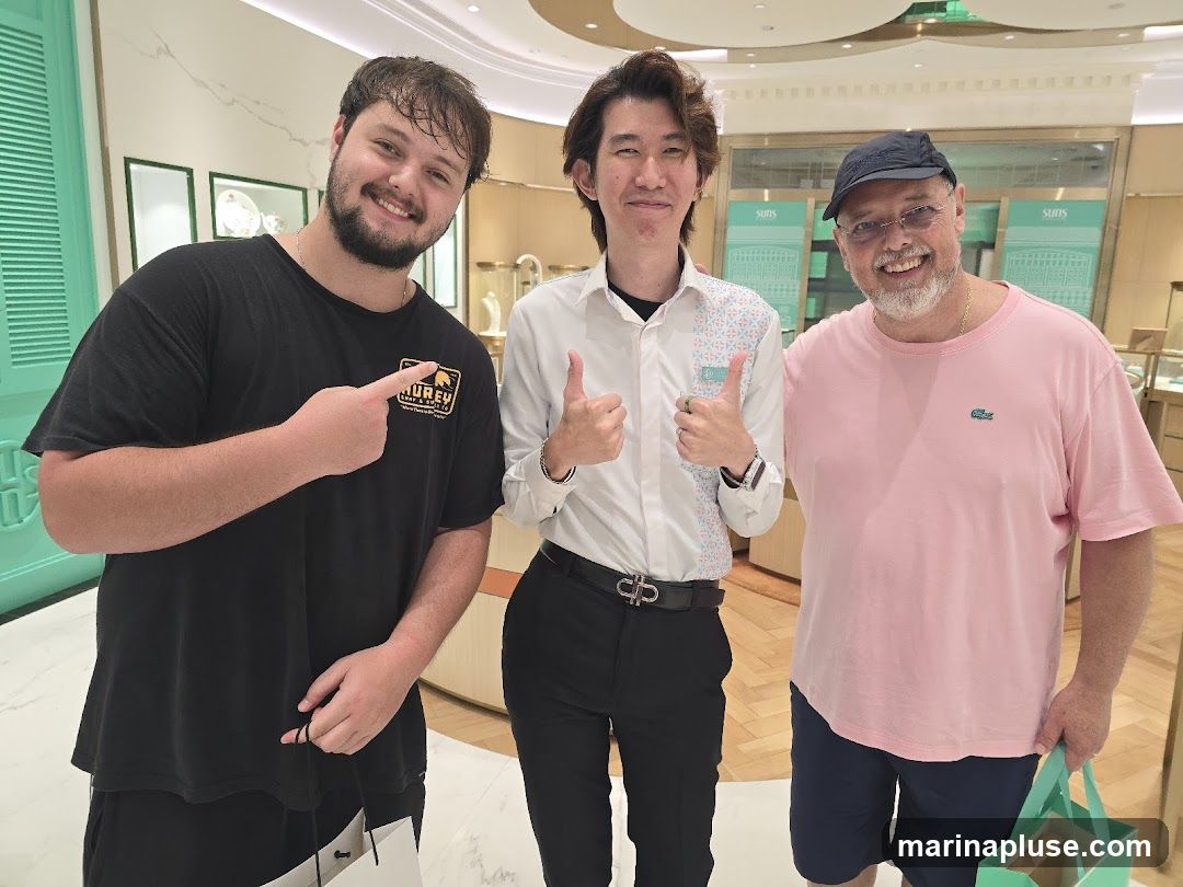 Three men are posing for a photo inside a brightly lit store. The man on the left is wearing a black t-shirt and pointing to his shirt. The man in the middle is wearing a white collared shirt with a patterned design and giving a thumbs up. The man on the right is wearing a pink t-shirt and a baseball cap, also giving a thumbs up. The background shows display cases with jewelry and the store's branding.