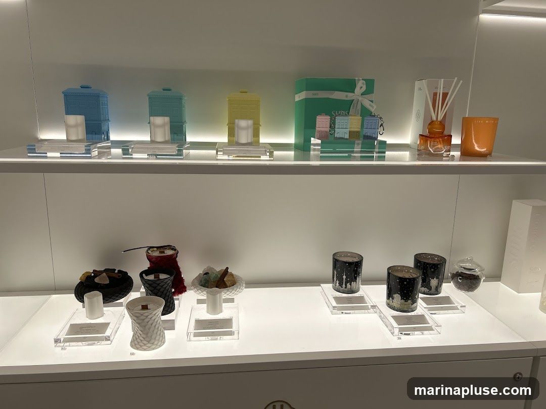 A retail display featuring various candles and home fragrance items on white shelves. The top shelf showcases decorative building-shaped candle holders in blue, teal, and yellow, along with a gift box, a diffuser with reeds, and an orange glass candle. The bottom shelf displays an assortment of candles in textured vessels, some adorned with decorative elements like crystals and cinnamon sticks, and others with cityscape designs. A small glass jar filled with coffee beans is also visible.