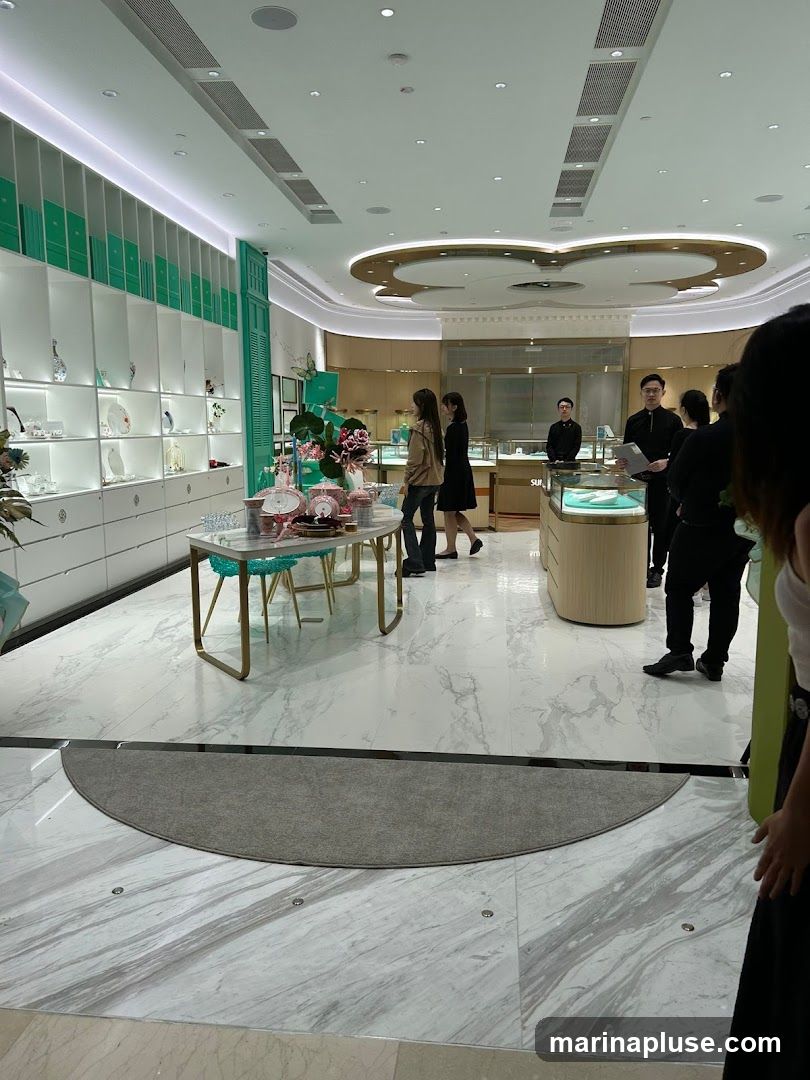 A well-lit jewelry store interior with display shelves filled with merchandise. Several customers and staff members are present, some browsing and others attending to customers. The store features a modern design with marble flooring and elegant display cases.