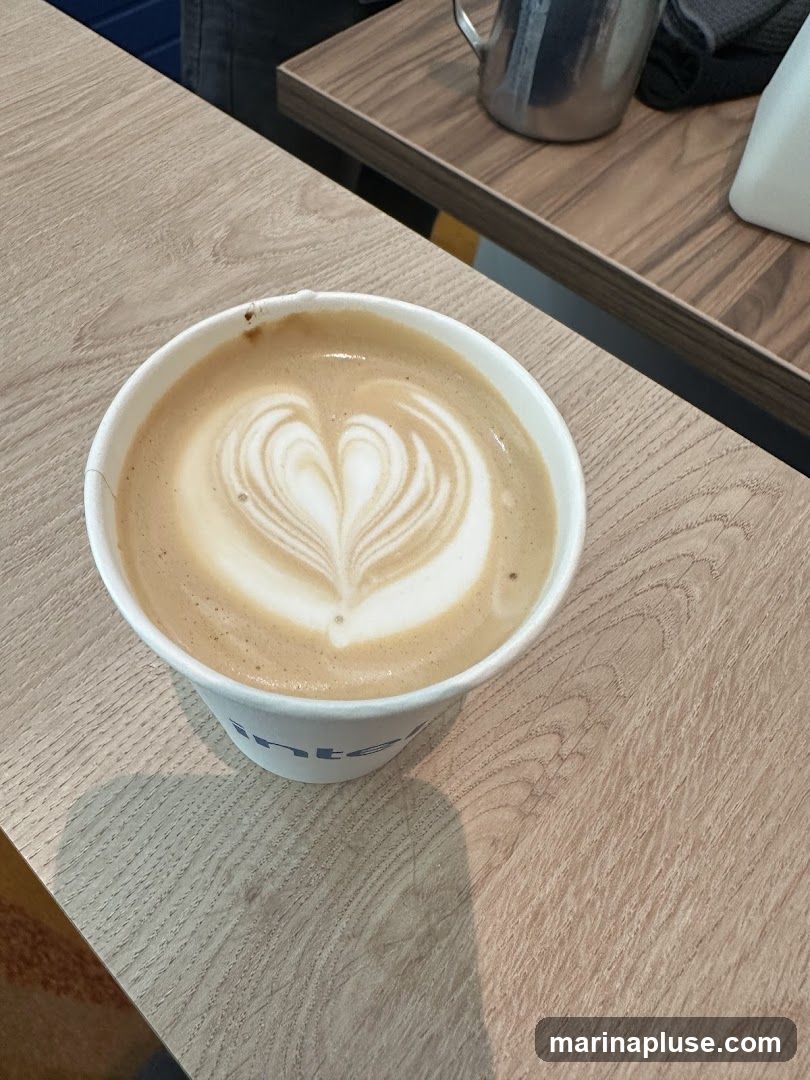 A close-up shot of a latte in a white paper cup, featuring intricate latte art in the shape of a heart. The cup has a blue logo visible on its side. The coffee is placed on a light-colored wooden surface, with a darker wooden shelf and a metal pitcher in the background.