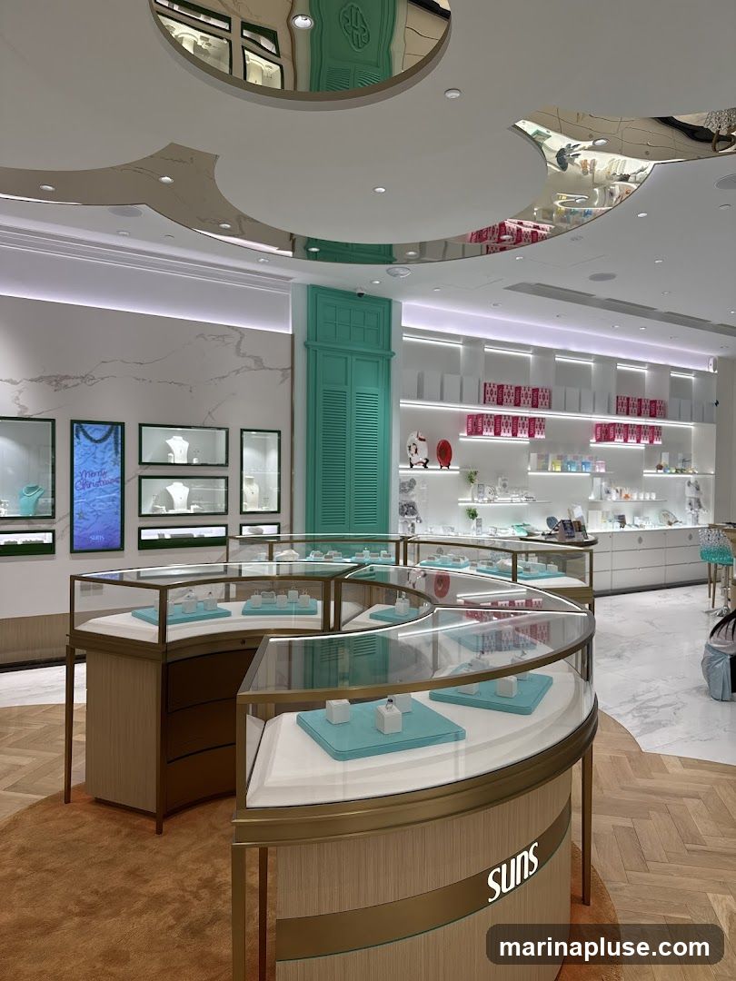A well-lit jewelry store interior with curved display cases showcasing rings and necklaces. The walls are adorned with marble-like panels and shelves stocked with various products. A distinctive teal-colored shuttered door is visible in the background. The ceiling features reflective panels that mirror the store's interior. The brand name 'SUNS' is visible on one of the display cases.