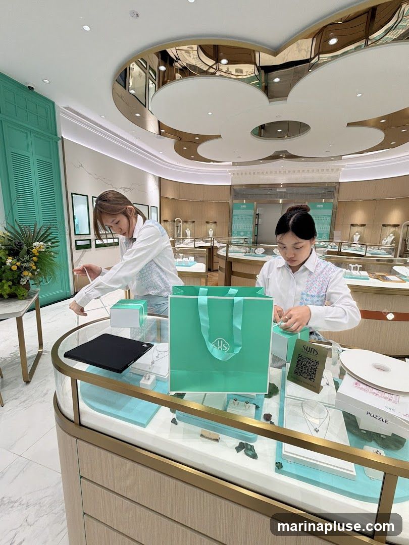 Two female employees are seen working in a brightly lit jewelry store. The store features display cases filled with jewelry, and a decorative ceiling with reflective panels. One employee is arranging items on a counter, while the other is packing a gift bag. A large teal shopping bag with a logo is prominent on the counter.