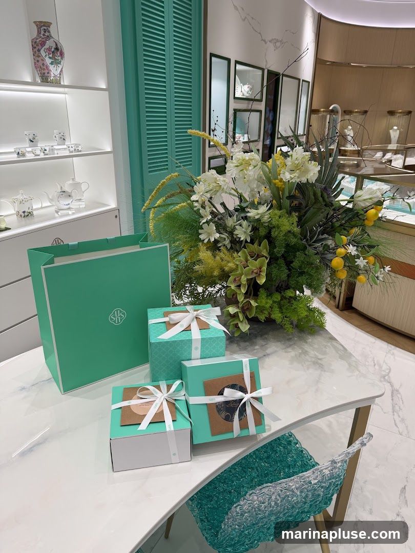 A display of gift-wrapped boxes and a large floral arrangement on a marble table in what appears to be a retail store. The boxes are teal and brown, tied with white ribbon. A teal shopping bag with a logo is also present. In the background, shelves display porcelain items, and a jewelry display case is visible. A decorative chair with a textured teal and white seat is in the foreground.