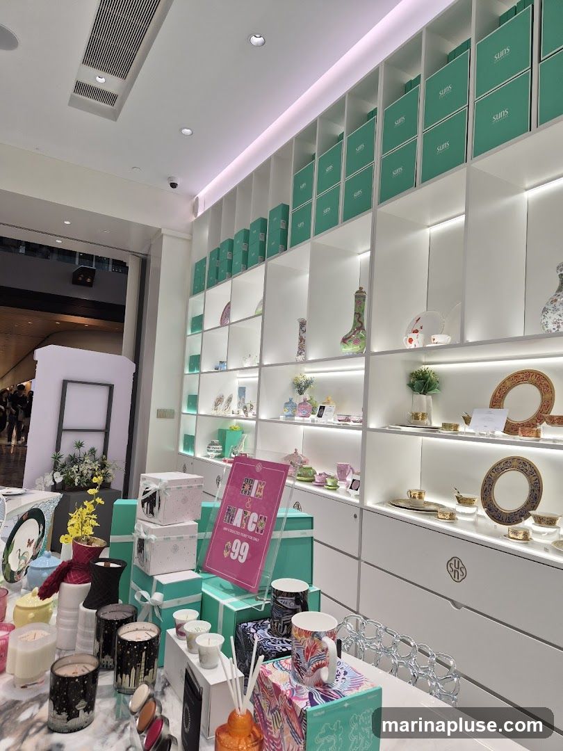 A brightly lit store interior showcasing a variety of decorative items on shelves and a display counter. The shelves are filled with teal boxes, ceramic plates, cups, and vases. The counter displays candles, diffusers, mugs, and gift boxes. A pink promotional sign reads "MIX & MATCH ANY 3 SELECTED ITEMS FOR $99".