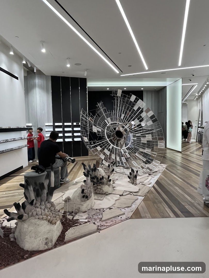 The image shows the interior of a modern retail space, possibly an optical store, with displays of eyewear on the left wall. In the center, a large, intricate metallic sculpture resembling a spiral or a fan is displayed on a platform with abstract, organic-shaped sculptures in the foreground. Several people are browsing or working in the store. The flooring is made of polished wood planks arranged in a diagonal pattern. The ceiling features sleek, linear lighting.