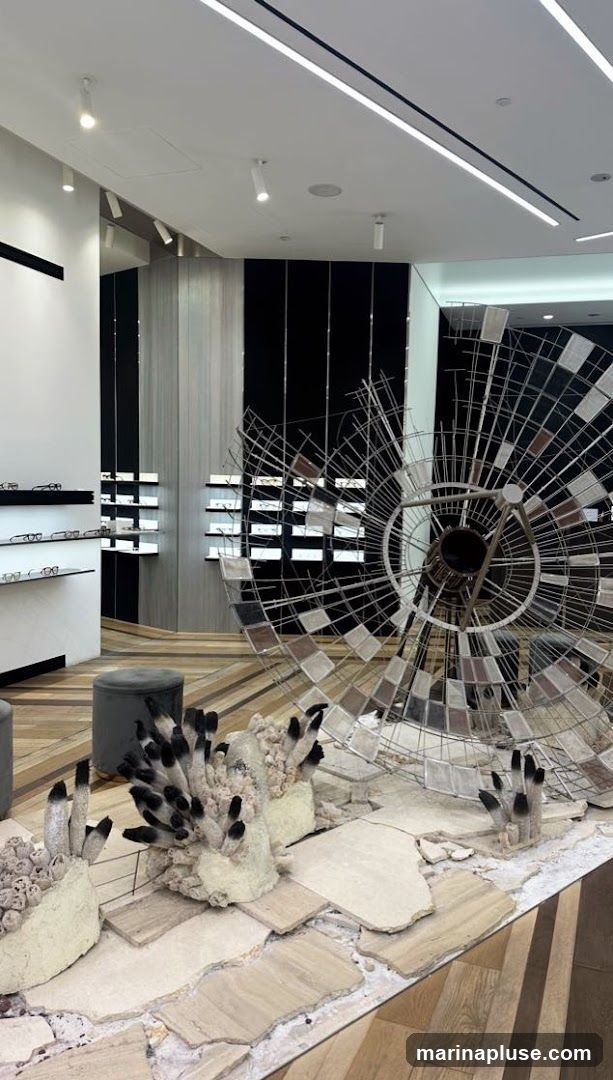 The interior of a modern eyewear store features sleek displays of eyeglasses on shelves. A large, abstract metal sculpture with reflective panels is a prominent decorative element in the foreground, alongside a display of artistic, coral-like sculptures. The flooring is a mix of wood planks arranged in a herringbone pattern and stone tiles.