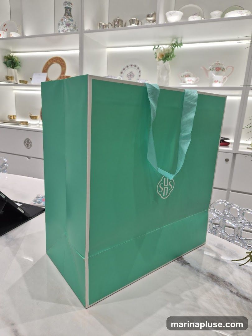 A teal gift bag with white trim and handles sits on a marble surface. A white logo is visible on the front of the bag. In the background, shelves display various porcelain items, including teacups, plates, and vases.