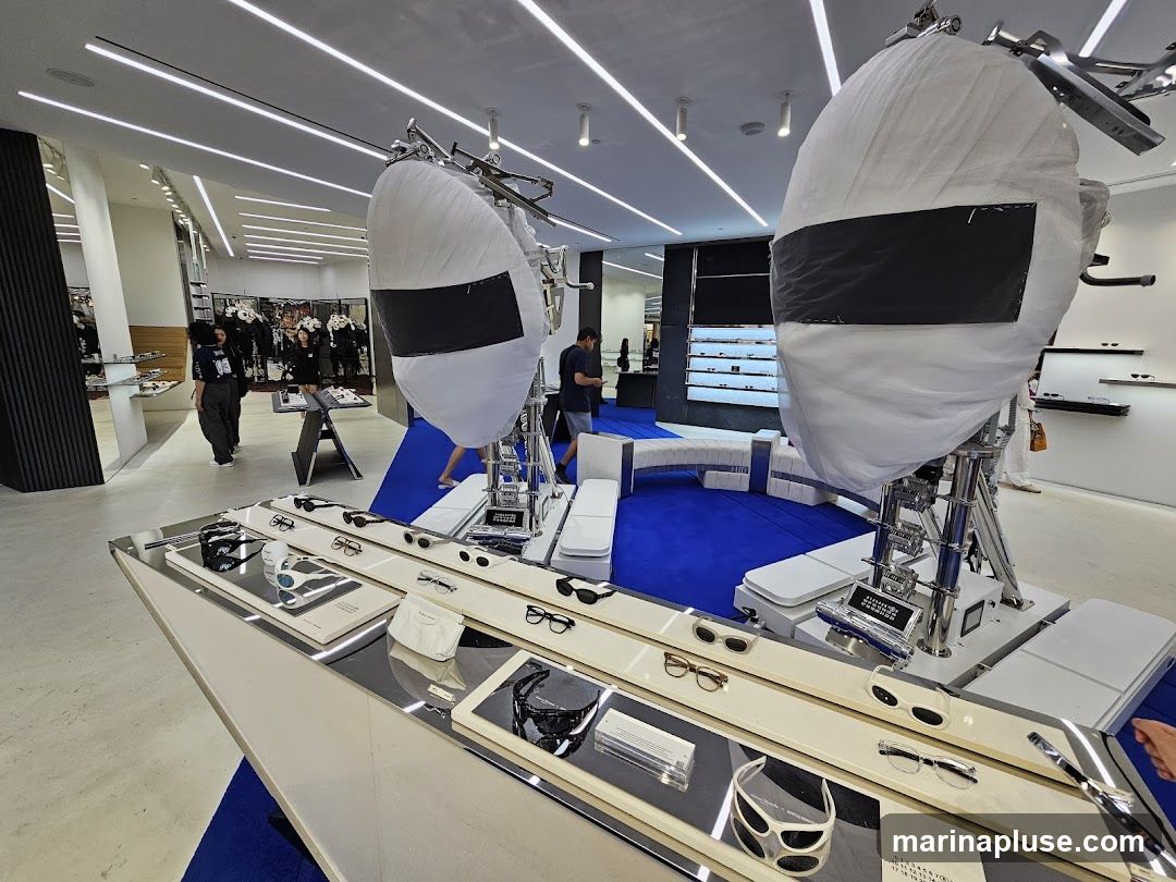 A modern eyewear store interior with displays of sunglasses and eyeglasses. In the foreground, a long, sleek display case showcases various sunglasses and eyeglasses on white stands. Behind this, two large, white, balloon-like structures covered in protective sheeting are prominent. Further back, more eyewear is displayed on shelves along the walls, and customers are browsing. The store features bright, linear ceiling lights and a vibrant blue carpet in the central area.