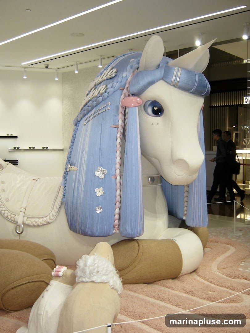 A large, whimsical unicorn sculpture with blue braided hair and pink accents is the centerpiece of a retail display. The unicorn is adorned with decorative buttons and a saddle. In the foreground, smaller plush figures resembling dogs or sheep are visible. The background shows a retail environment with shelves and people walking by.
