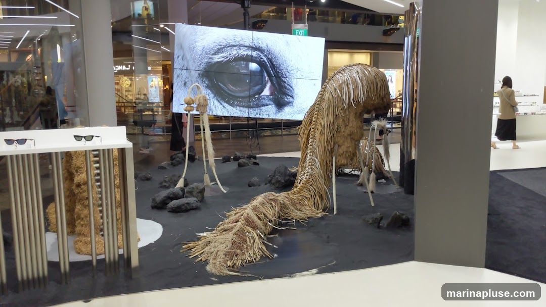 An art installation features a large screen displaying a close-up of a horse's eye. In front of the screen, a sculpture resembling a creature with a long, fringed tail made of straw and wood is displayed on a black surface with rocks. To the left, a display stand holds two pairs of sunglasses. In the background, a person walks past a store with shelves of products.