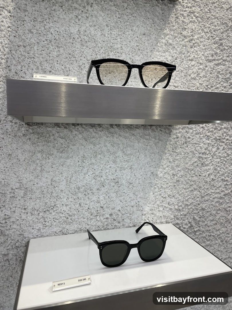Two pairs of black-framed sunglasses are displayed on shelves against a textured white wall. The top pair has a slightly more angular frame and lighter lenses, while the bottom pair has a rounder, thicker frame and darker lenses. Small labels with product names and prices are placed in front of each pair.