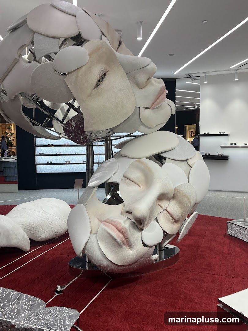 A large, abstract sculpture resembling multiple faces is displayed prominently on a red carpet in what appears to be a retail environment. The sculpture is made of various materials, including what looks like plaster or resin, with delicate features and layered elements. In the background, shelves display eyewear, suggesting the store might be an optician or a high-end fashion retailer. The lighting is bright and modern, with linear ceiling lights.
