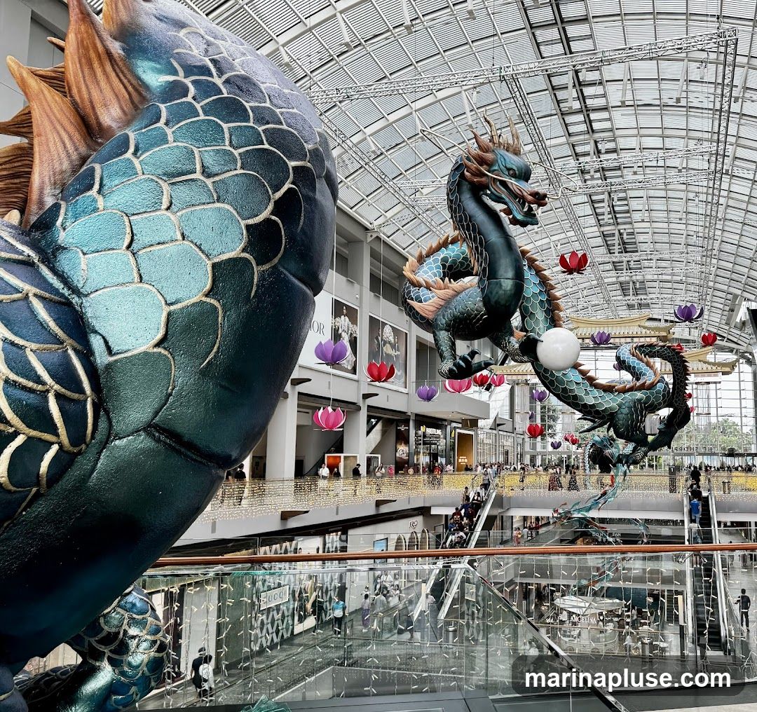 A large, detailed dragon sculpture is prominently displayed in the foreground, with its scales shimmering in shades of teal and gold. In the background, several other dragon sculptures are suspended from the ceiling of a modern shopping mall, which features a glass roof and multiple levels. Colorful lotus-shaped lanterns in pink, red, and purple hang among the dragons. People can be seen browsing and walking on escalators and walkways on different floors of the mall. The overall scene suggests a festive or celebratory atmosphere, possibly for a holiday like Lunar New Year.
