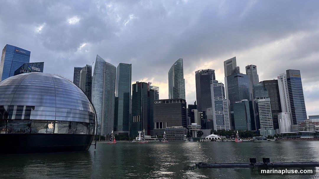 A panoramic view of the Singapore skyline at dusk, featuring modern skyscrapers and a distinctive spherical building on the left. The water in the foreground reflects the cloudy sky and the city lights. Several sailboats with red sails are visible on the water. Buildings include HSBC, Standard Chartered, and others with visible signage. The spherical building appears to be an Apple Store, with its logo visible.