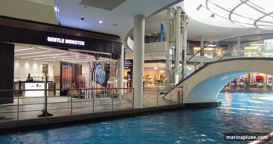 The exterior of a Gentle Monster store is visible, with its name illuminated above the entrance. Inside, displays of eyewear and decorative items can be seen. A person wearing a mask is standing behind a counter. The store is located within a modern shopping mall with a canal-like water feature in the foreground and escalators and other shops in the background.