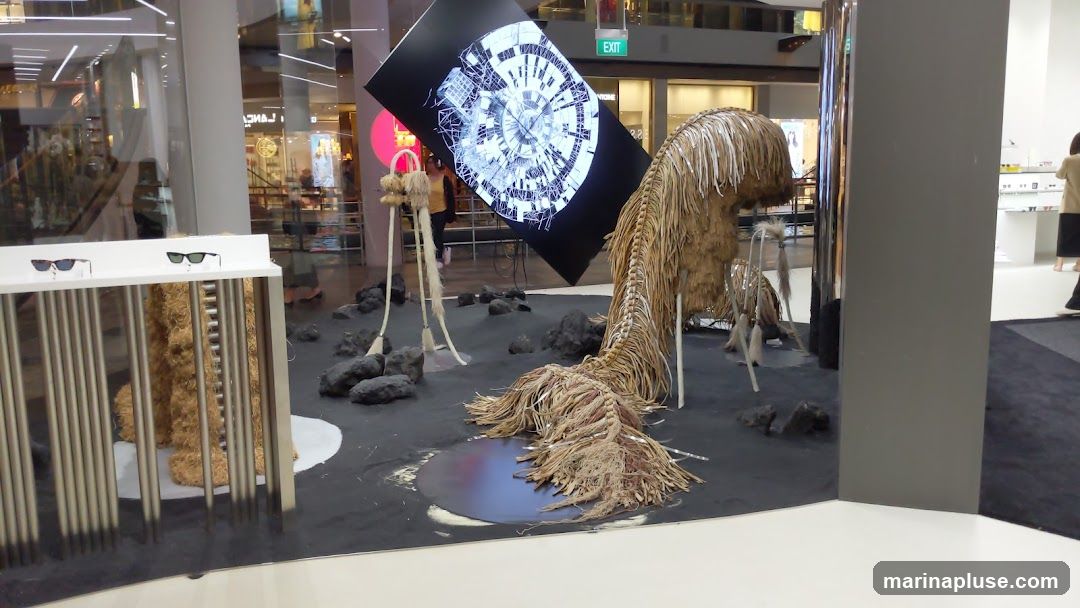 An artistic display features a large, abstract sculpture resembling a creature with a long, flowing tail made of natural fibers. In front of the sculpture, a white display stand showcases two pairs of sunglasses. Behind the sculpture, a large digital screen displays a complex, circular graphic. The display is set against a dark, textured surface resembling sand or earth, with scattered rocks. In the background, reflections of a shopping mall interior are visible, including escalators and shopfronts. To the right, a display of eyewear is visible, suggesting the context of an optical store.
