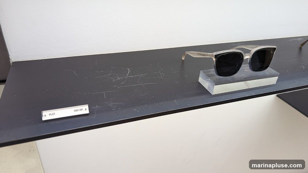A pair of light-colored sunglasses with dark lenses is displayed on a clear acrylic stand on a dark shelf. A small white label with the text "PLOT SGD 381" is visible on the shelf in front of the sunglasses. The background is a plain white wall.