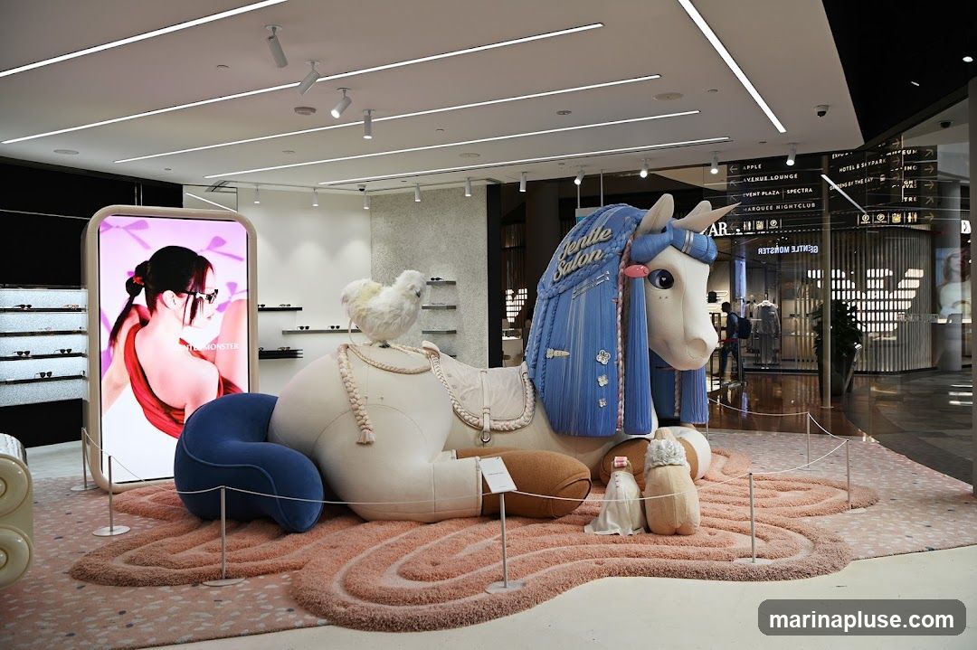 A large, plush unicorn display with a white chicken on its back is set up in a store. The unicorn has blue hair and is adorned with various accessories. A large digital screen displays an advertisement for Gentle Monster eyewear. In the background, store shelves with glasses are visible, along with signage for other businesses in the mall.