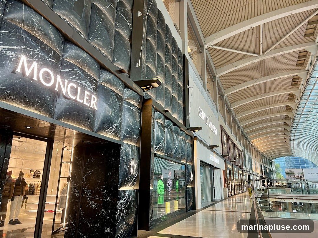The exterior of a Moncler store with its distinctive marble-patterned facade and illuminated logo. The entrance is visible, showcasing mannequins dressed in the brand's apparel. Adjacent to it, other luxury brand stores like Gentle Monster are also visible along the indoor shopping mall corridor.