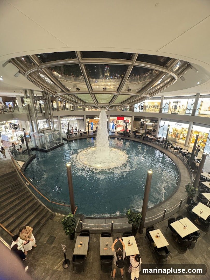 An indoor view of a large circular fountain with water jets, surrounded by multiple levels of shops and restaurants. Several tables and chairs are set up around the fountain area, with people seated and walking around. The ceiling features a glass dome reflecting the interior lights and activity.