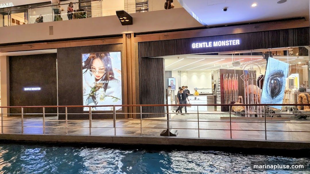 The exterior of a Gentle Monster store is visible, featuring large illuminated displays and the store's name prominently displayed. The store has a modern facade with glass windows and dark paneling. Inside, displays of eyewear and artistic installations can be seen. People are walking in and out of the store. The store is situated along a waterway.