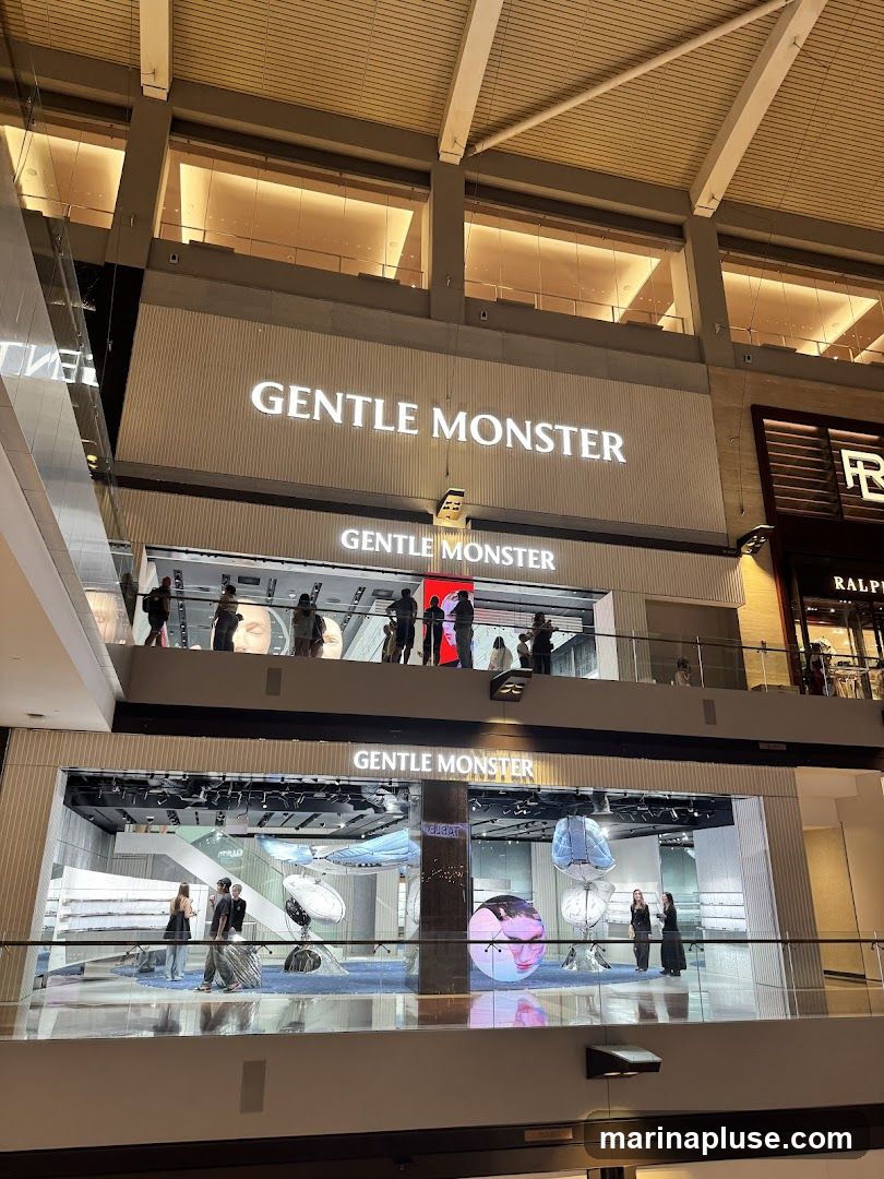 A multi-level store with the brand name "GENTLE MONSTER" displayed prominently on its facade. The lower level features a large glass storefront showcasing an interior with artistic displays and people browsing. The upper level also has a visible storefront with people standing on a balcony.
