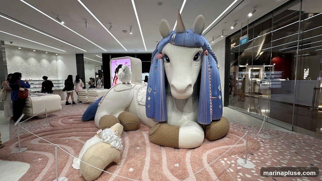 A large, plush unicorn with blue braided hair and a small white bird perched on its back is the centerpiece of a retail display. A smaller plush sheep is positioned in front of the unicorn. The display is set on a pink rug with a wavy pattern. In the background, there are shelves with products, possibly eyewear, and a glass storefront with an "EXIT" sign. Several people are browsing in the store.