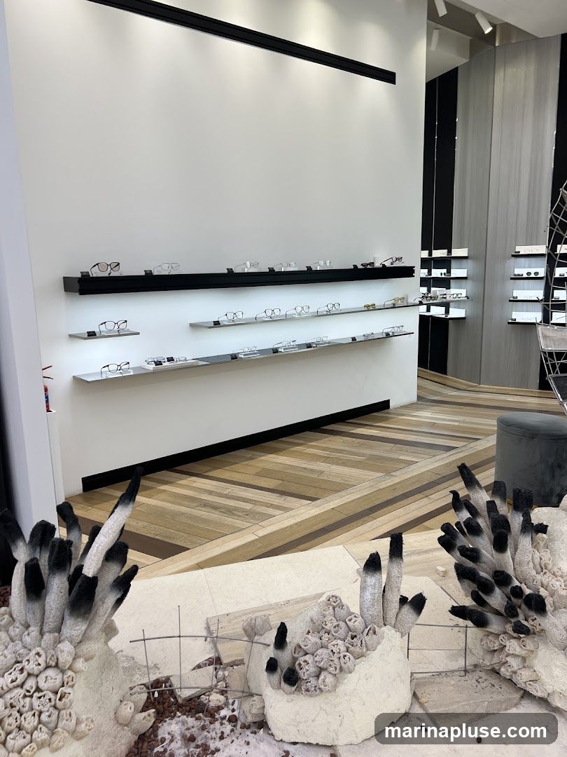 A modern retail space with shelves displaying various eyeglasses. The walls are white, and the flooring is a light wood parquet. In the foreground, there are abstract sculptural elements resembling fungi or coral, made of white and black materials.