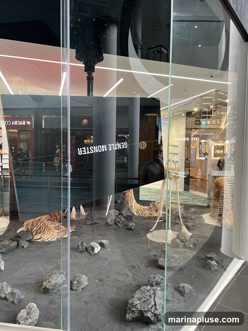 A storefront display features an abstract art installation with black sand, rocks, and dried plant material. The reflection in the glass shows the interior of a shopping mall with various shops and signage. The name "GENTLE MONSTER" is visible on a display inside the store.