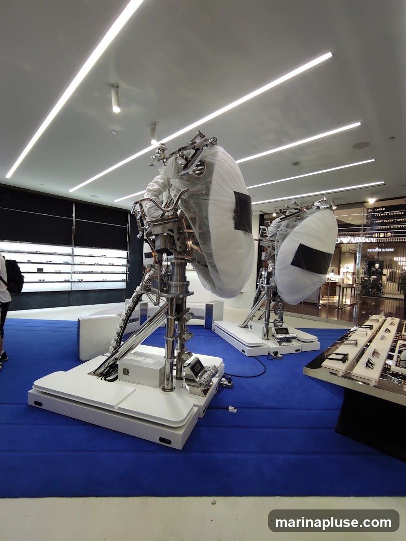Two large, metallic structures covered in protective plastic are displayed on white platforms on a blue carpet. The structures appear to be part of an exhibition or display. In the background, there are shelves with products and signage for various shops, including "ARMANI EXCHANGE". A display case with watches is visible to the right.