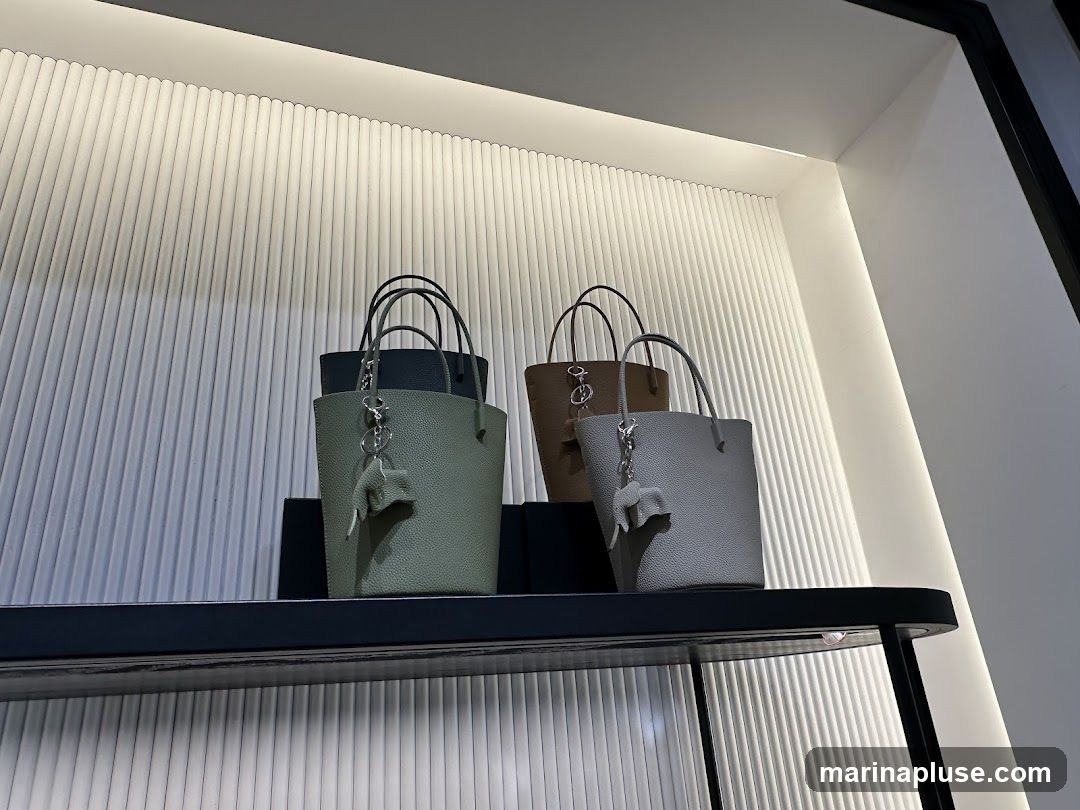 Four textured handbags are displayed on a shelf in a store. The handbags are in shades of sage green, dark blue, brown, and light grey. Each handbag has a top handle and a small elephant-shaped charm attached to the handle. The background features a ribbed white wall with integrated lighting.