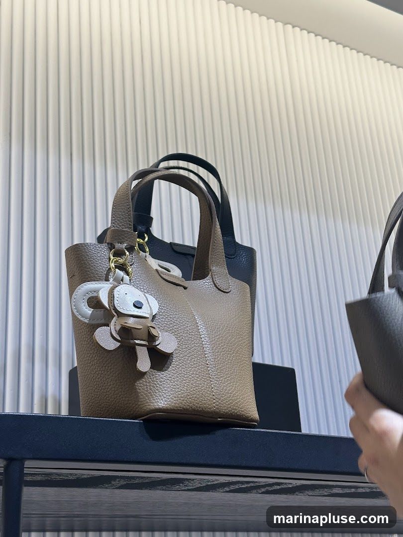 A close-up shot of several textured leather handbags displayed on a shelf. The handbag in the foreground is a light brown color and features a playful koala-shaped charm attached to its handle. Behind it, a black handbag is visible, and to the right, a hand reaches for a dark gray handbag. The background has a ribbed texture.
