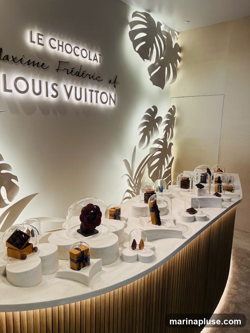 A display of chocolates and confections presented in a luxurious setting. The wall behind features illuminated text reading "LE CHOCOLAT Maxime Frédéric at LOUIS VUITTON" and decorative cutouts of monstera leaves. The chocolates are arranged on a curved counter with various pedestals and covered by glass domes.