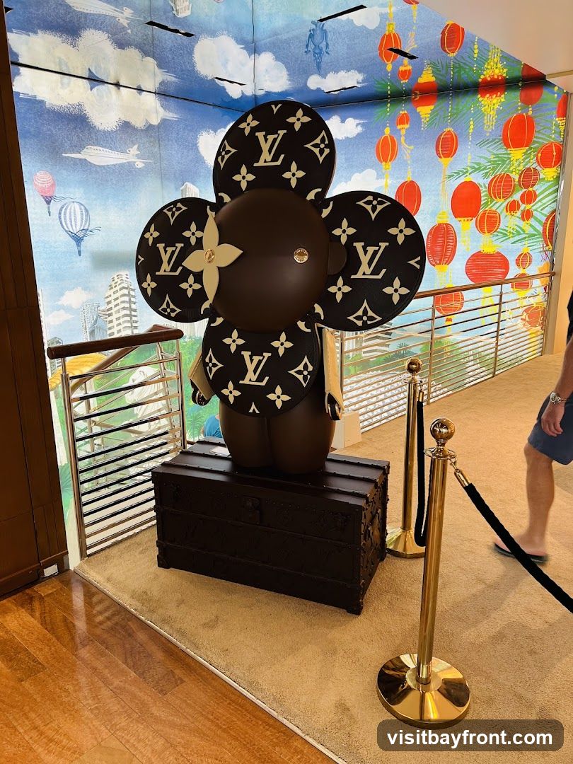 A large, dark brown, cartoon-like figure with flower-shaped ears and chest adorned with the Louis Vuitton monogram pattern stands on top of a dark brown Louis Vuitton trunk. The background features a mural with a sky scene, hot air balloons, airplanes, and city buildings on the left, and a scene with red lanterns and yellow foliage on the right. Gold stanchions with black ropes are in the foreground. A person wearing shorts and a t-shirt is walking in the background on the right.