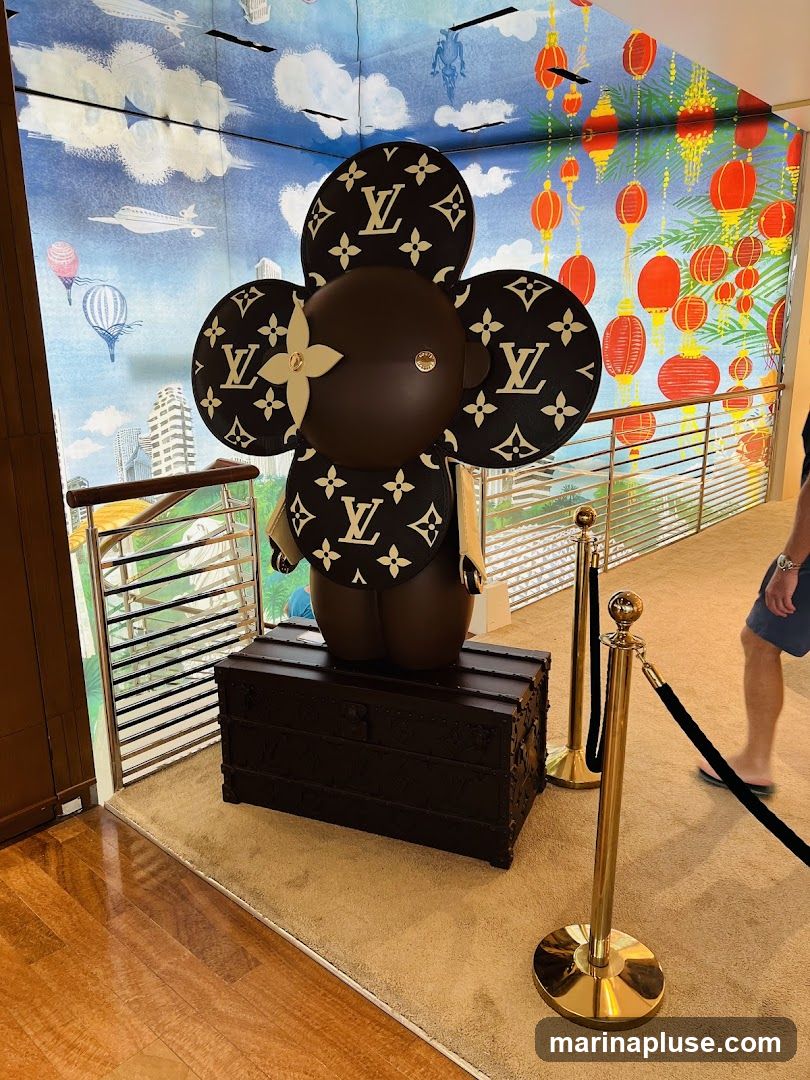 A large, dark brown, cartoon-like figure with flower-shaped ears and chest adorned with the Louis Vuitton monogram pattern stands on top of a dark brown Louis Vuitton trunk. The background features a mural with a sky scene, hot air balloons, airplanes, and city buildings on the left, and a scene with red lanterns and yellow foliage on the right. Gold stanchions with black ropes are in the foreground. A person wearing shorts and a t-shirt is walking in the background on the right.