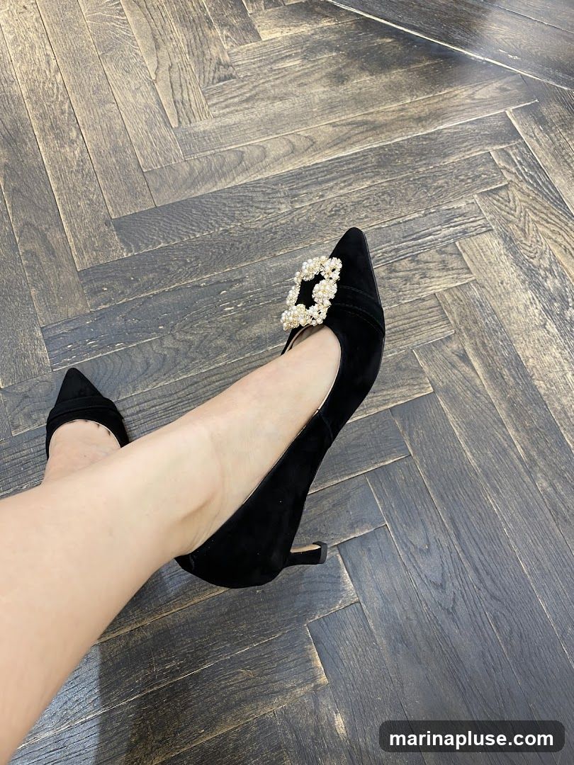 A close-up shot of a person's legs and feet wearing black suede pointed-toe heels. The shoes feature a decorative buckle adorned with pearls and rhinestones on the vamp. The background is a dark, herringbone-patterned wooden floor.