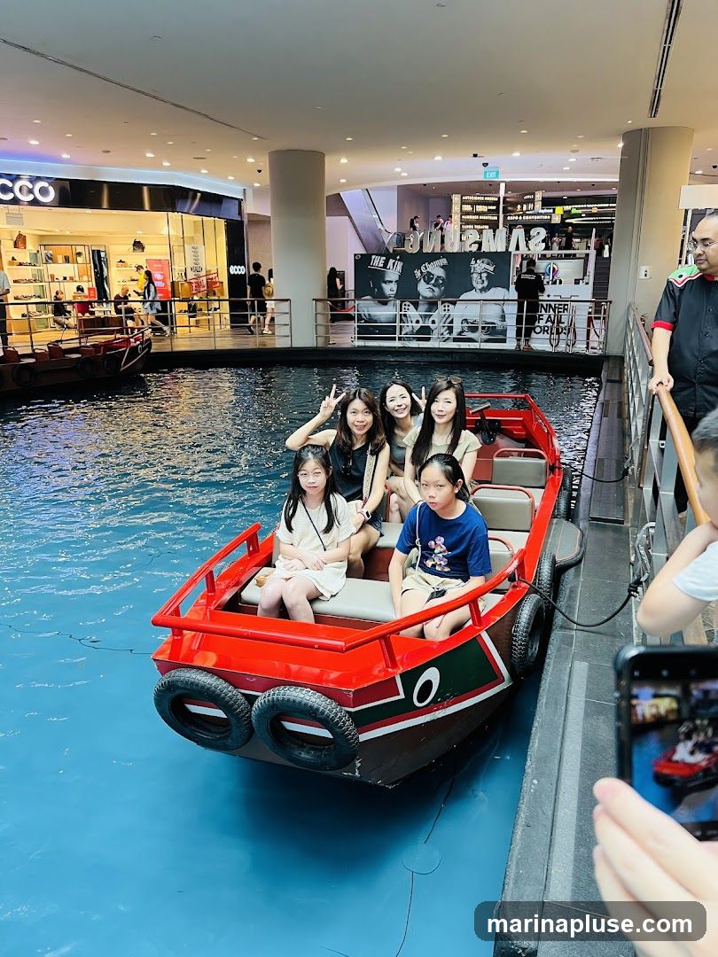 A group of people, including children and adults, are enjoying a boat ride on a canal-like waterway inside a shopping mall. The boat is red and green with black tires as fenders. In the background, there are shops with visible signage, including "ECCO". There are also escalators and other shoppers. One person is taking a photo of the boat with their phone.