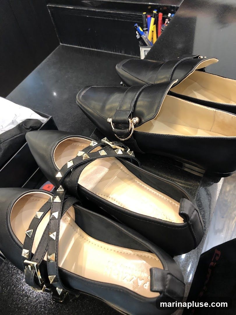 A close-up shot of two pairs of black flat shoes. The pair in the foreground has multiple straps adorned with gold studs and buckles. The pair behind it is a loafer style with a gold ring detail. The shoes are displayed on a dark surface, possibly a counter.