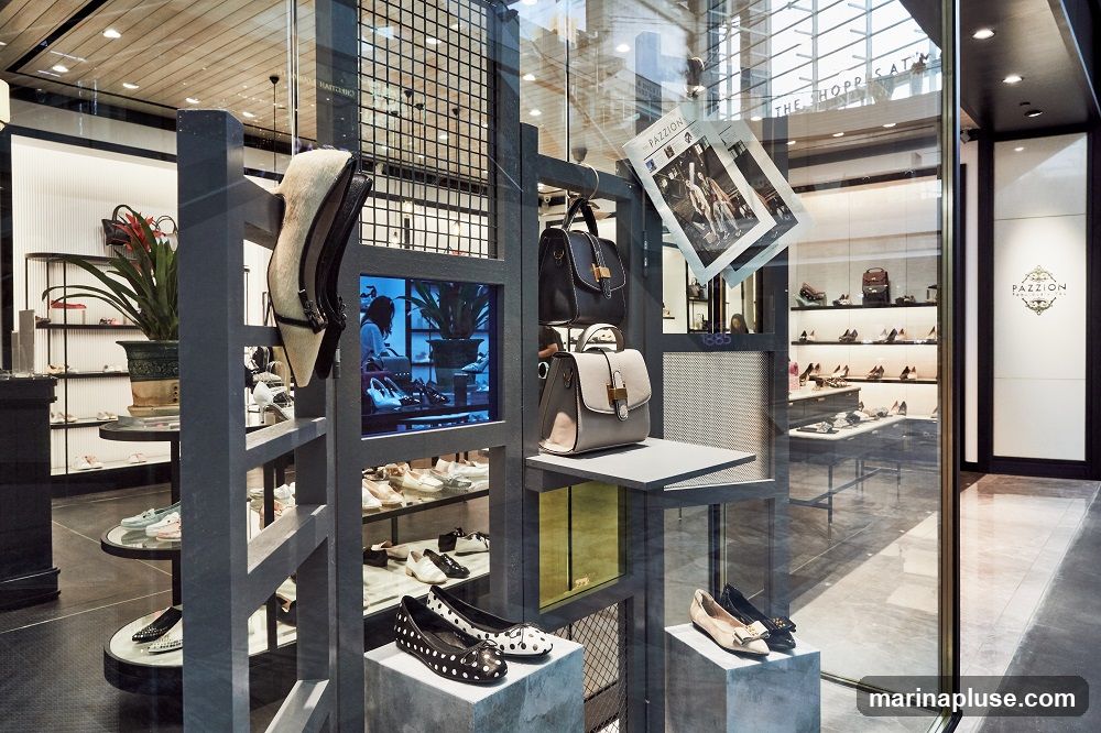 A well-lit retail display showcasing a variety of women's shoes and handbags. The display features a modern, industrial-chic aesthetic with grey shelving units and metal accents. Shoes are arranged on shelves and pedestals, with some displayed prominently in the foreground. Handbags are hung and placed on shelves. A digital screen within the display shows a fashion-related scene. In the background, more shoes are visible on shelves, and a sign with the brand name "PAZZION" is displayed on the right wall.